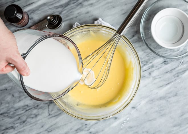 Añadiendo leche a la mezcla de crema pastelera en un recipiente hondo de vidrio y con un batidor de globo