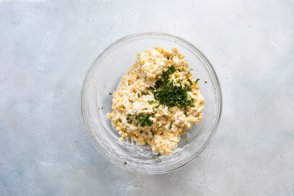 Granos de elote, mayonesa y queso rallado mezclados con cilantro en un recipiente hondo de vidrio